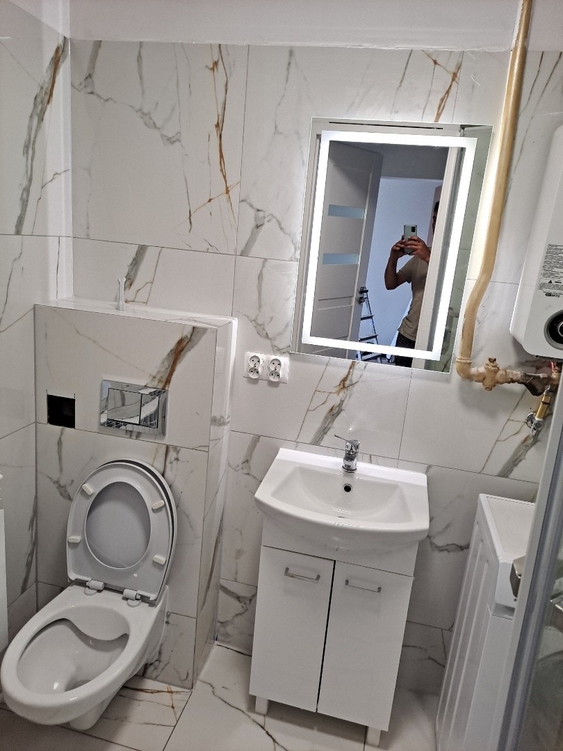 Wykończona łazienka z marmurowymi płytkami, białą umywalką z szafką, toaletą i podświetlanym lustrem, w którym odbija się osoba robiąca zdjęcie.