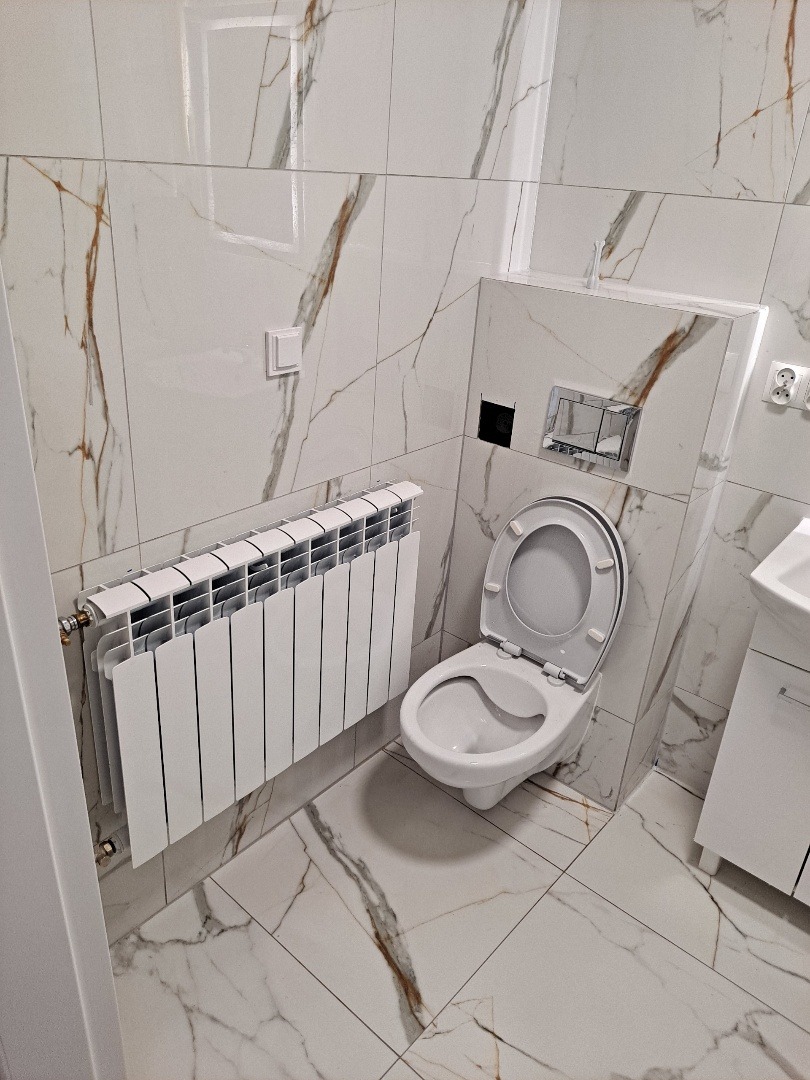 Biała łazienka z marmurowymi płytkami, wiszącą toaletą i grzejnikiem. Minimalistyczny design, jasne oświetlenie.