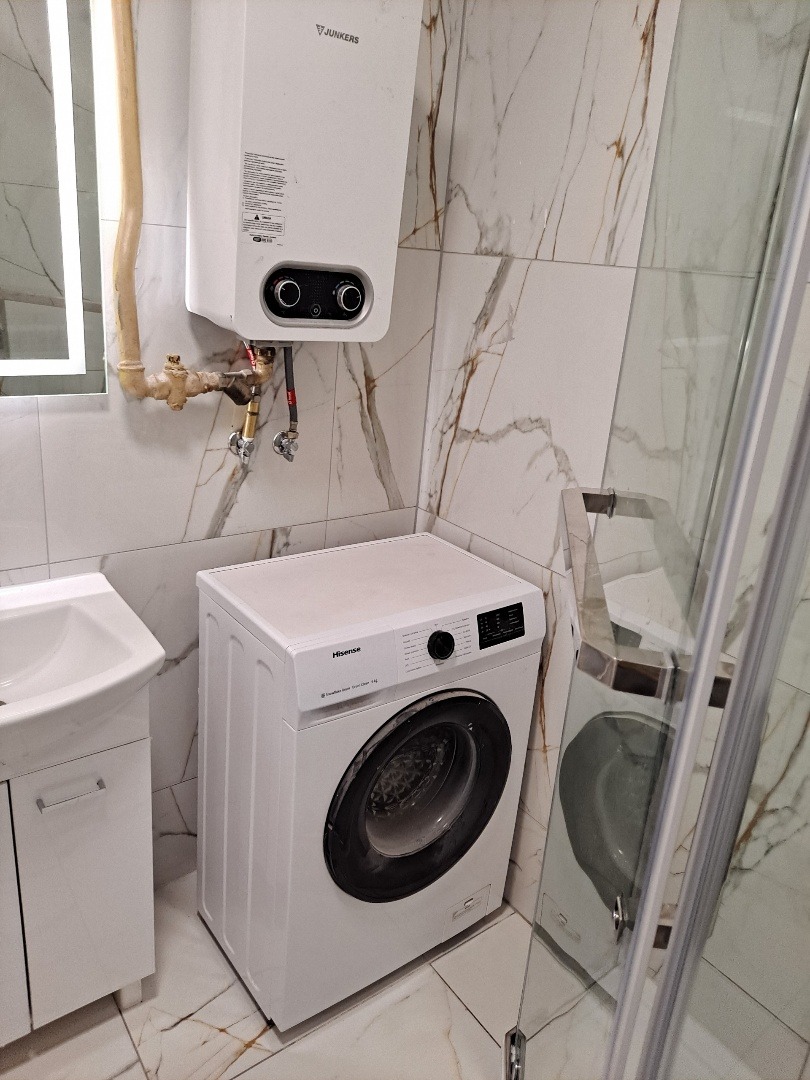 Wykończona łazienka z marmurowymi płytkami, białą pralką Hisense, umywalką z szafką i bojlerem Junkers. Kabina prysznicowa ze szklanymi drzwiami.