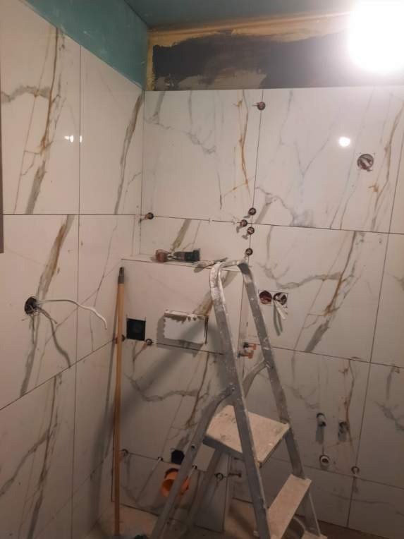 Łazienka w trakcie wykańczania: białe płytki imitujące marmur z brązowymi żyłami, drabina, przygotowane otwory pod armaturę. Etap prac wykończeniowych.