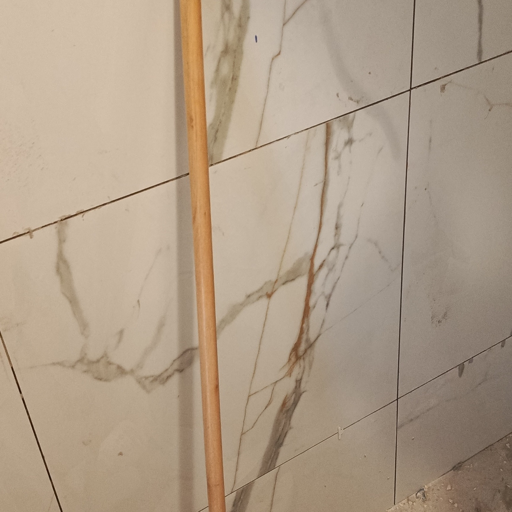 Ściana wyłożona białymi płytkami imitującymi marmur, z widocznym drewnianym narzędziem, prawdopodobnie podczas procesu wykańczania łazienki.