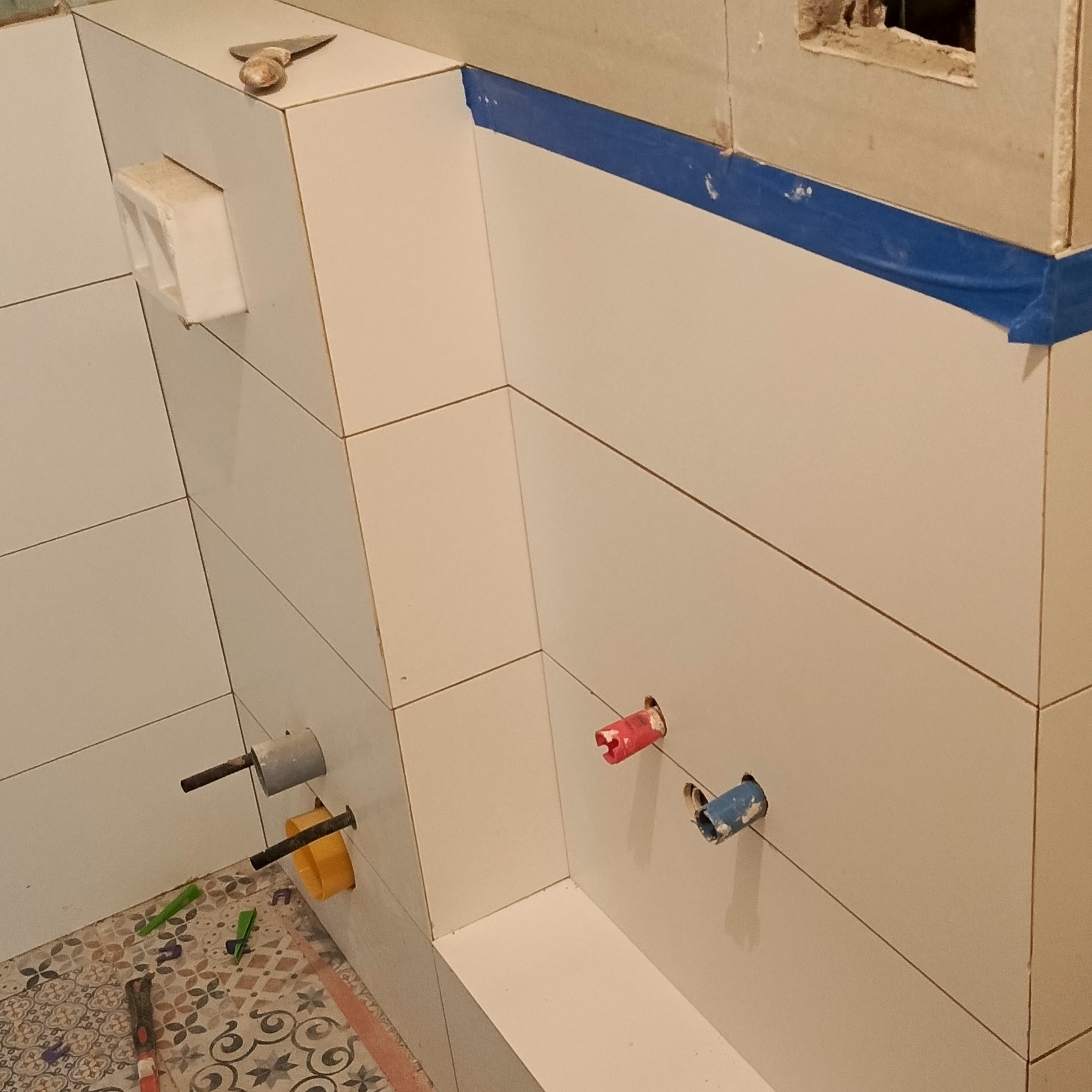 Układanie białych płytek w łazience. Widoczne kolorowe rury, narzędzia i niebieska taśma malarska. Prace wykończeniowe w toku.