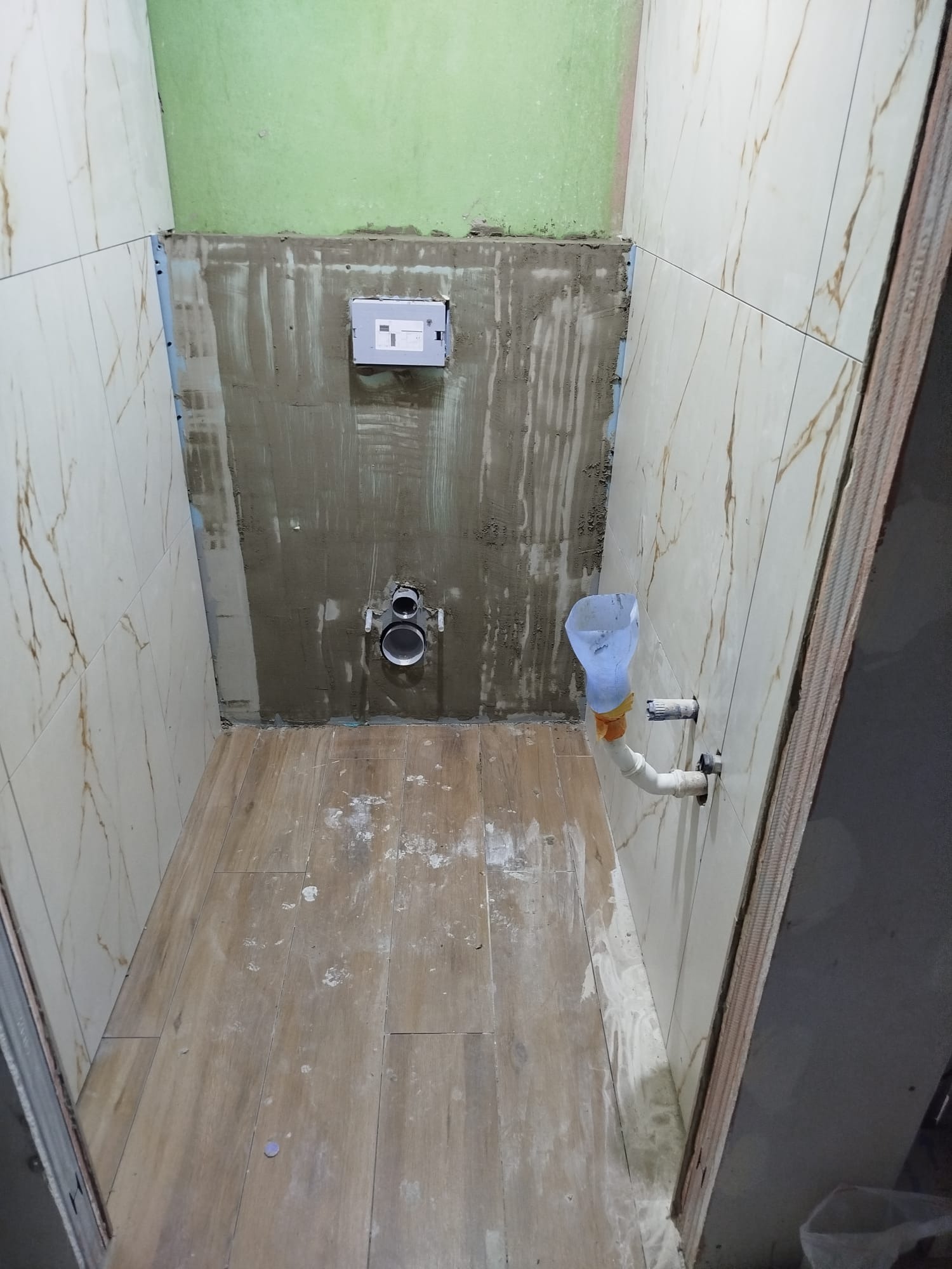 Remont łazienki w toku: ułożone płytki imitujące marmur na ścianach, drewnopodobne panele na podłodze, widoczna instalacja sanitarna i fragment zielonej ściany.