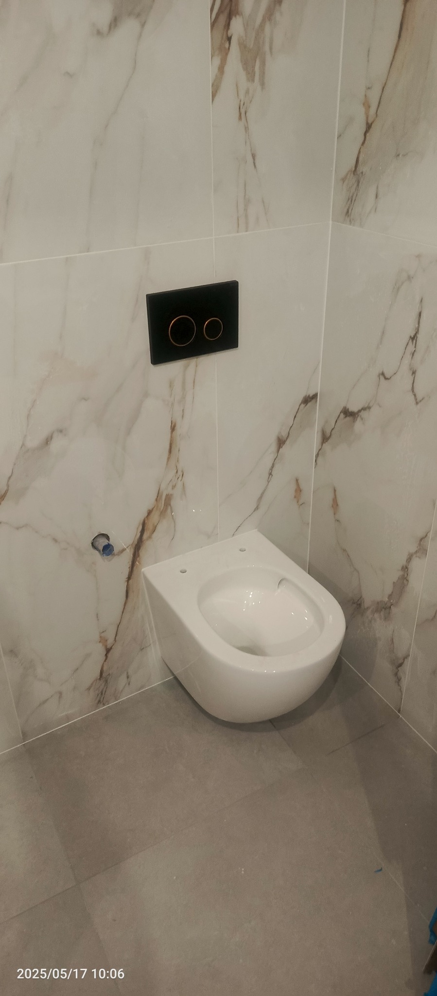 Wisząca miska WC z czarnym przyciskiem spłukującym na marmurowej ścianie w wykończonej łazience, szare płytki podłogowe. Minimalistyczny design.