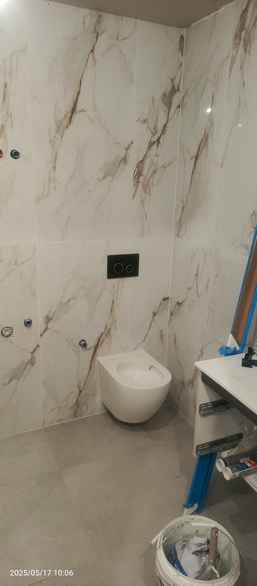 Bidet wiszący na ścianie z marmurowymi płytkami. Widoczne przygotowane otwory na baterię i przycisk spłukujący. Ukończona instalacja w łazience.