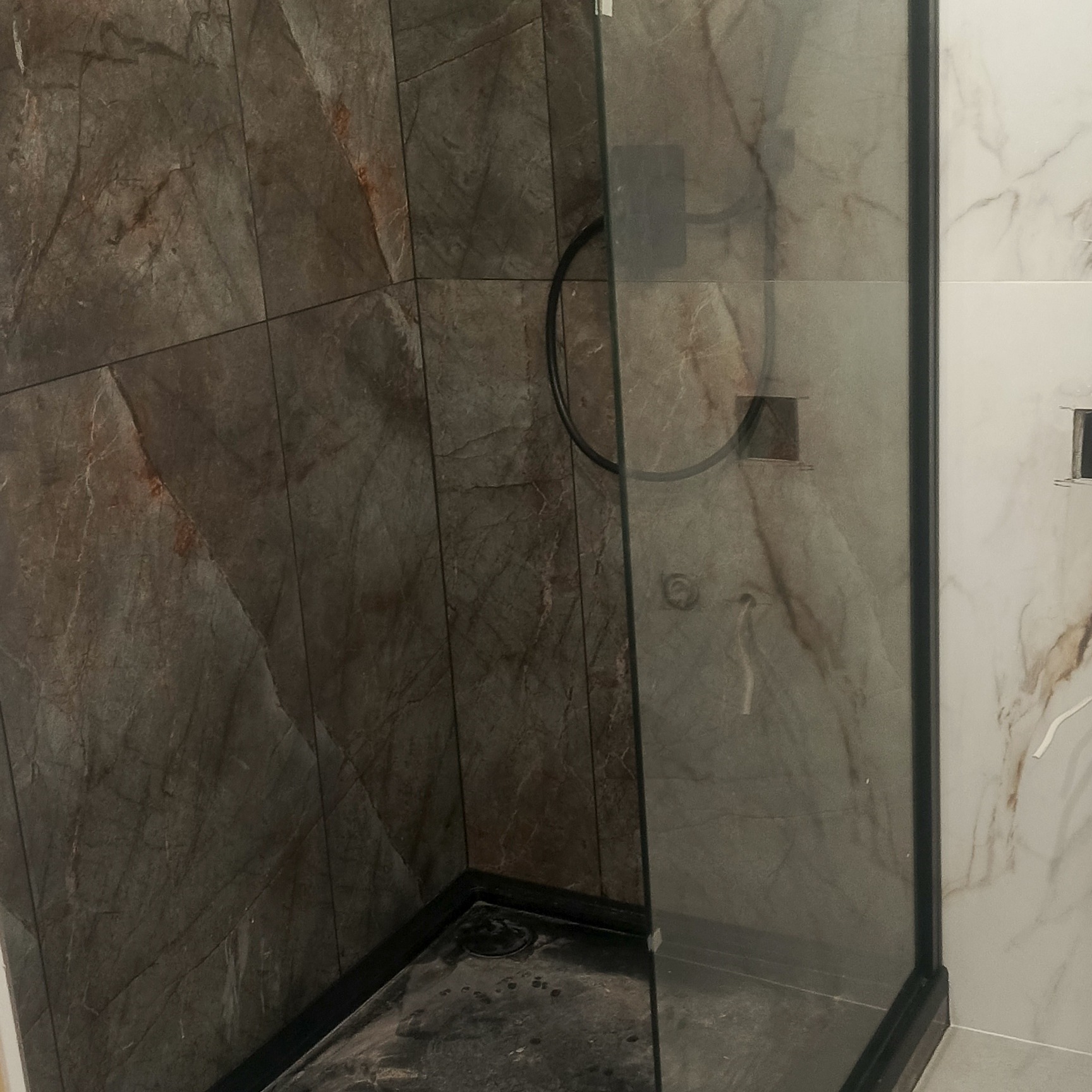 Nowoczesna kabina prysznicowa z ciemnymi, marmurkowymi płytkami i czarnymi detalami, widoczna na tle jasnych płytek.