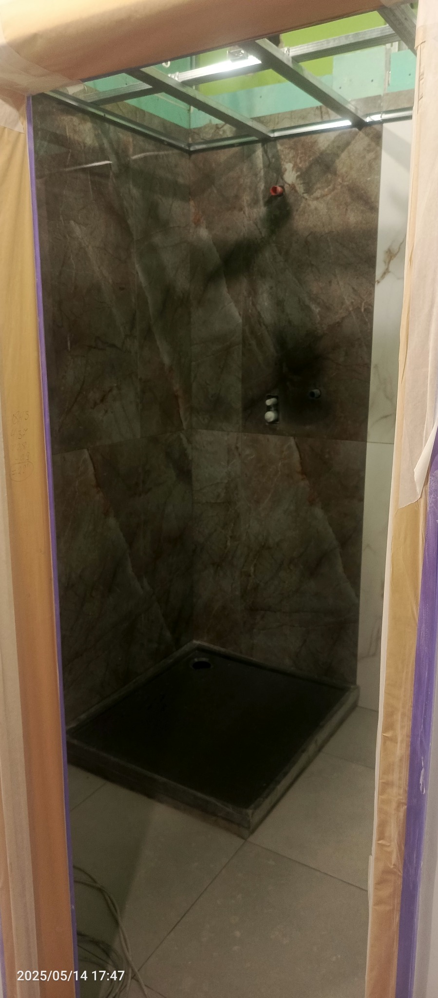 Wykończony brodzik z marmurowymi płytkami w kabinie prysznicowej, widoczne metalowe profile sufitowe i folia ochronna, co wskazuje na prace wykończeniowe w toku.