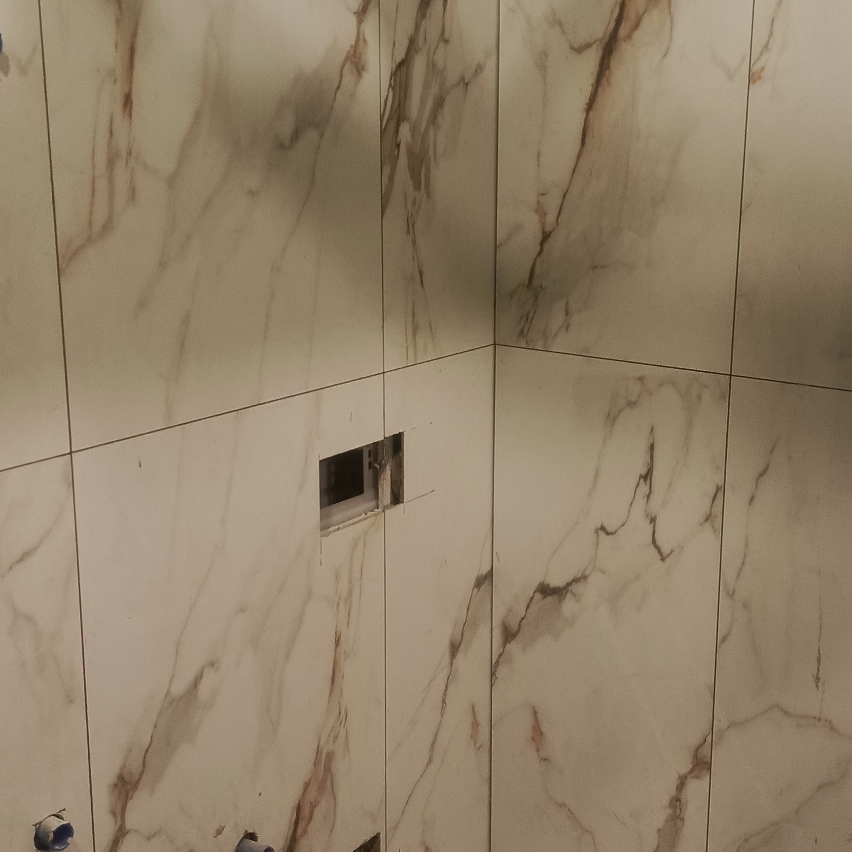 Ściana wyłożona białymi płytkami imitującymi marmur. Widoczny otwór na armaturę i fragment instalacji wodnej. Wykończenie łazienki w trakcie realizacji.