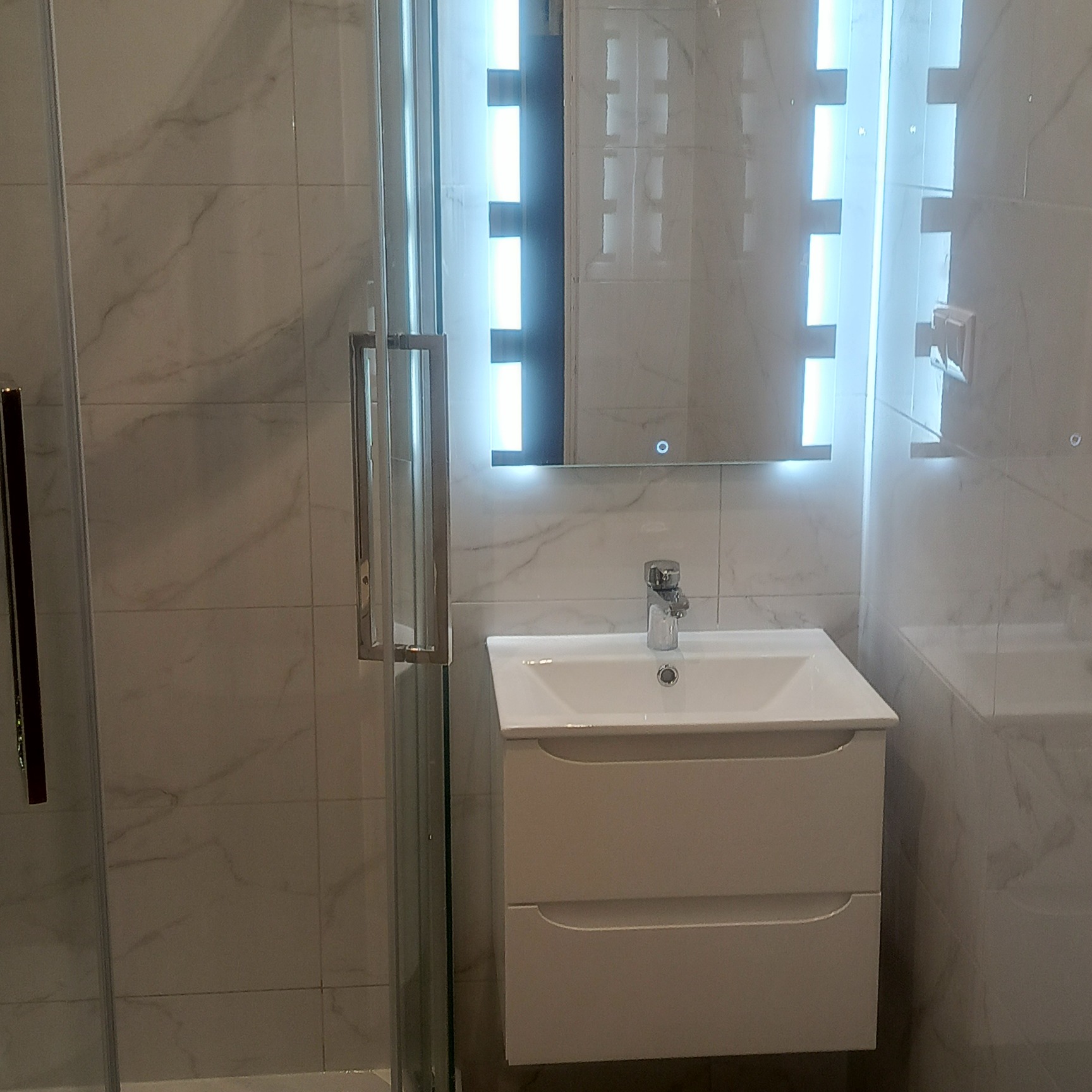 Nowoczesna łazienka z kabiną prysznicową, umywalką z szafką i lustrem z podświetleniem LED. Marmurowe płytki na ścianach.