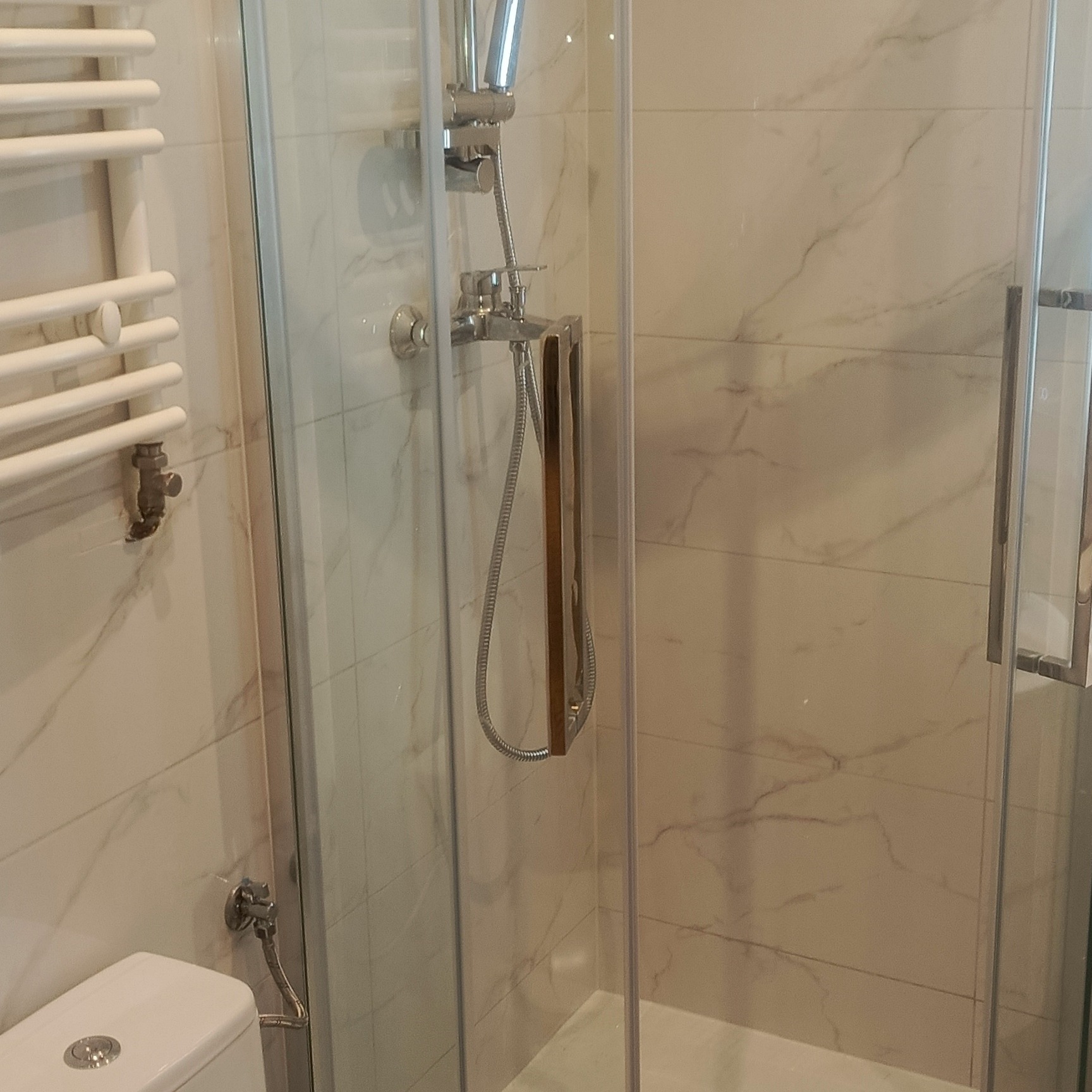 Nowoczesna kabina prysznicowa z marmurowymi płytkami, grzejnik łazienkowy i fragment toalety. Wykończenie łazienki w jasnych barwach, chromowane elementy.