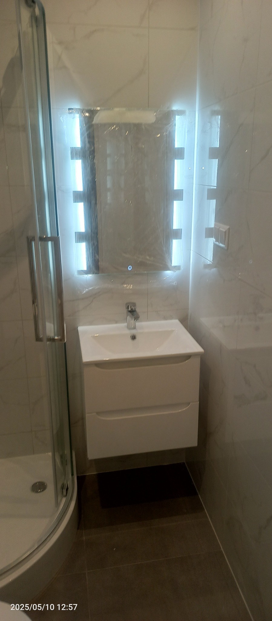 Nowoczesna łazienka z kabiną prysznicową, białą szafką z umywalką i podświetlanym lustrem, które jest jeszcze zabezpieczone folią ochronną.