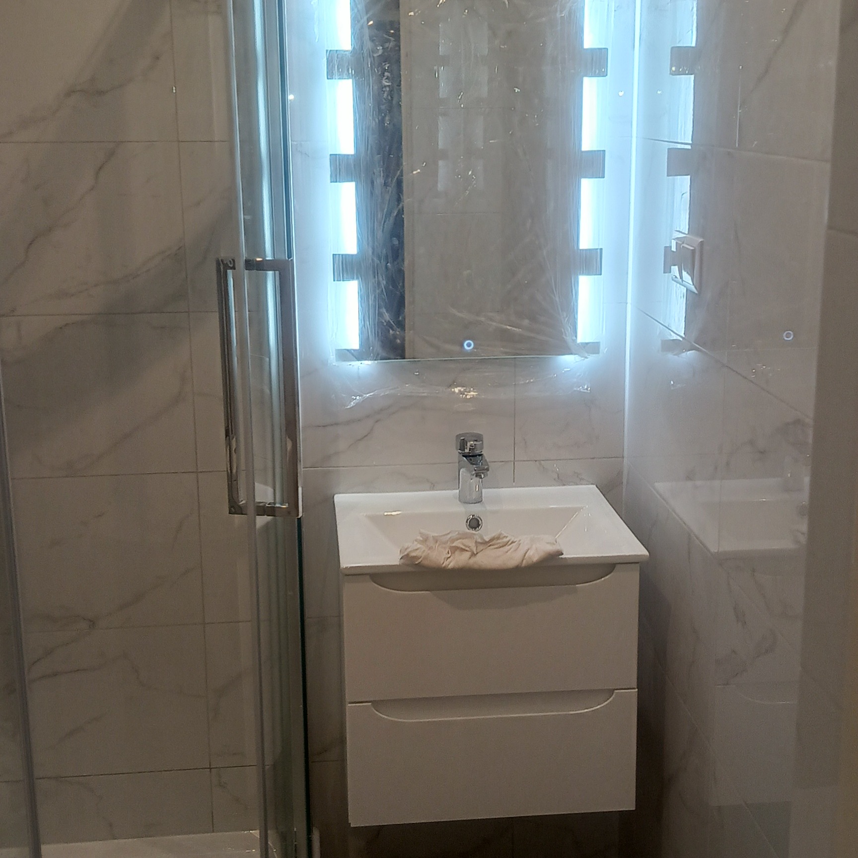 Nowoczesna łazienka: biała umywalka z szafką, podświetlane lustro z folią ochronną, marmurowe płytki. Widoczny fragment kabiny prysznicowej.