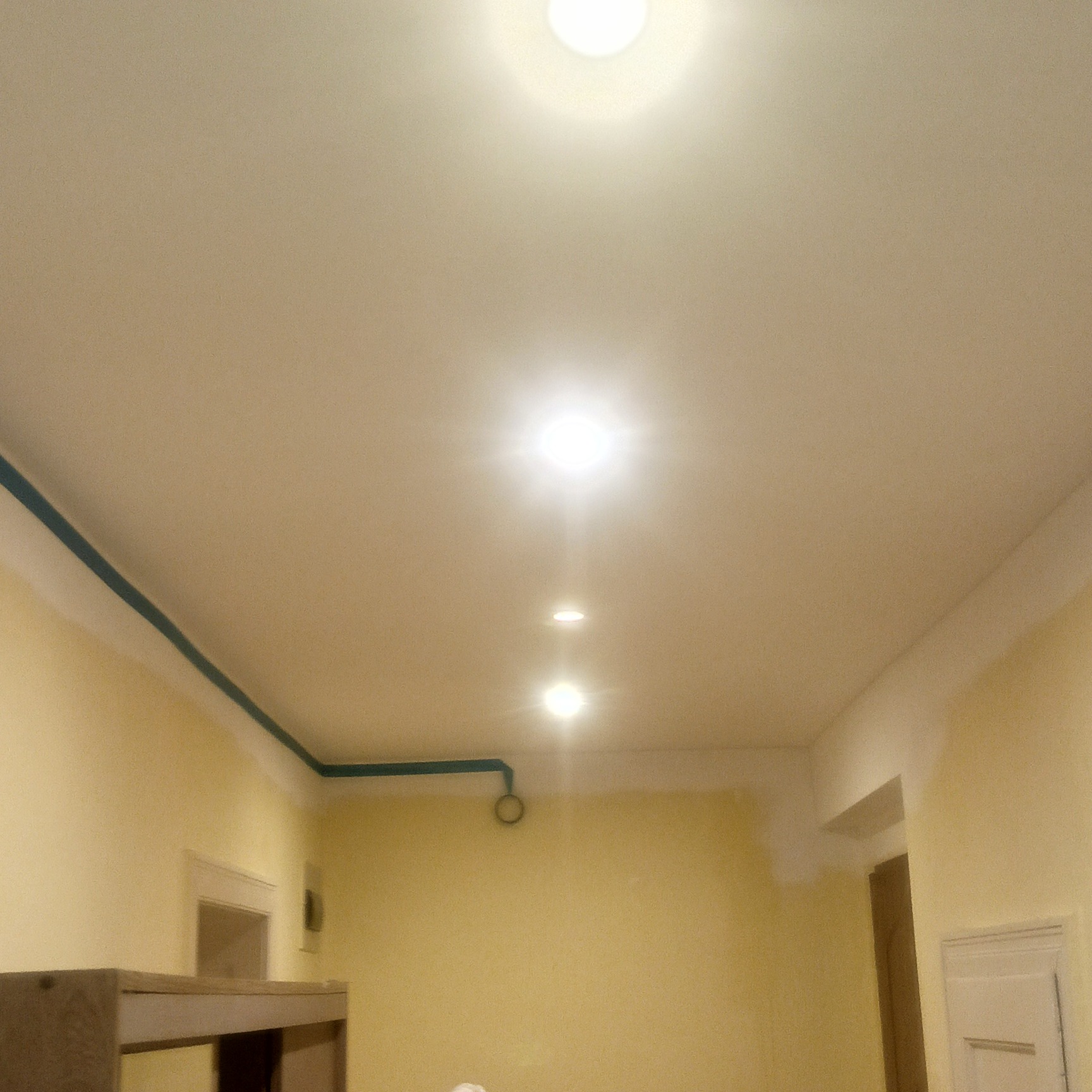 Wnętrze w trakcie malowania: żółte ściany, biały sufit z oświetleniem punktowym, widoczne niebieskie rury przy suficie. Częściowo widoczne meble.