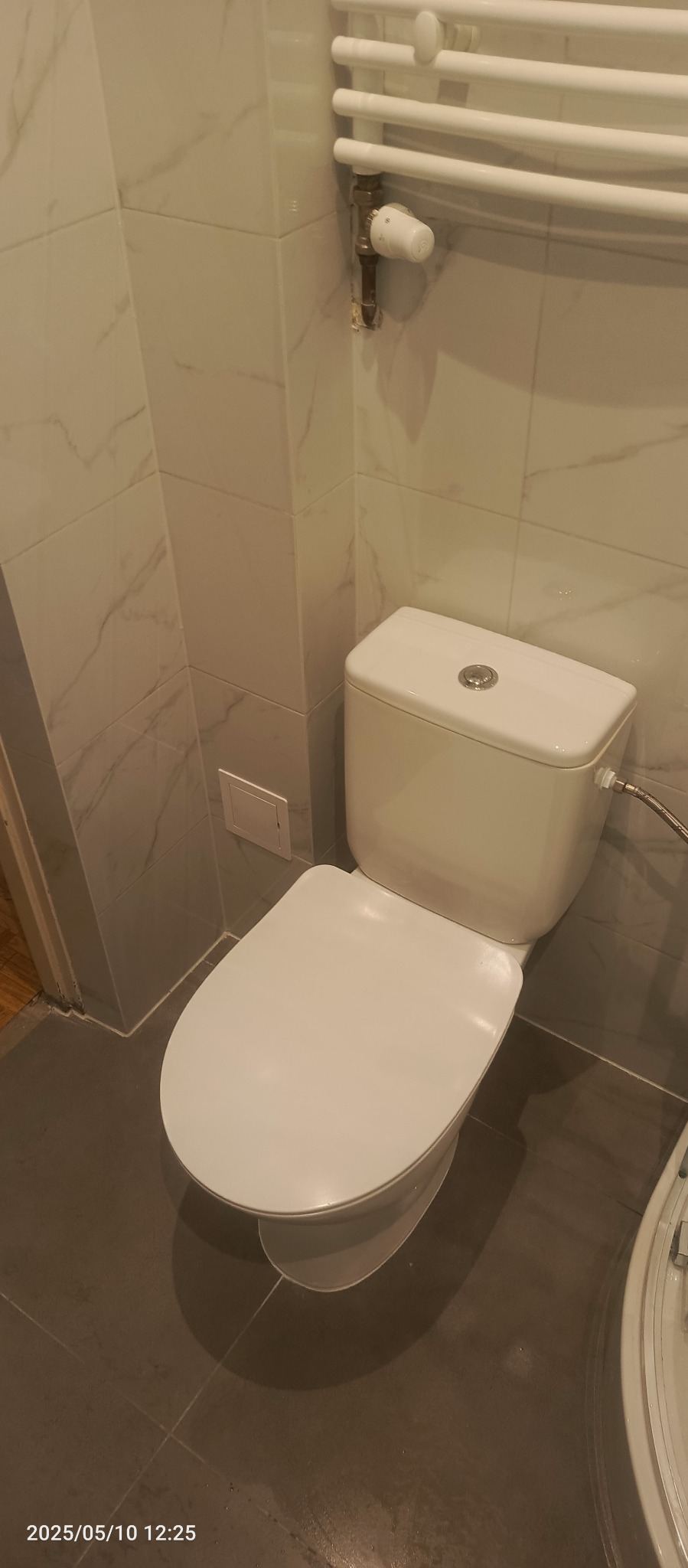 Biała toaleta w łazience z marmurowymi płytkami ściennymi i grzejnikiem drabinkowym. Widoczny fragment wanny. Nowoczesne wykończenie.