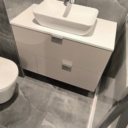 Lukasz Fabisiak - Beżowa szafka łazienkowa z dwiema szufladami i białą umywalką nablatową, chromowany kran, szare płytki podłogowe i ścienne z marmurkowym wzorem.