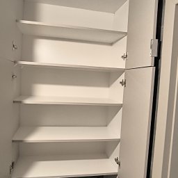 Lukasz Fabisiak - Otwarta, biała szafa z półkami, widok z bliska na wnętrze. Drzwiczki szafy częściowo otwarte, ukazujące metalowe zawiasy. Minimalistyczny design, jasne oświetlenie.