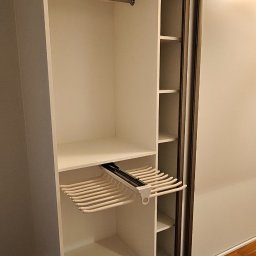 Lukasz Fabisiak - Biała szafa przesuwna z wysuwanym wieszakiem na spodnie i półkami, widok z bliska, oświetlenie sufitowe z wikliny, parkiet na podłodze.