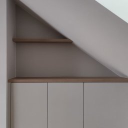 Lukasz Fabisiak - Szafka wnękowa z szarymi frontami i drewnianym blatem, dopasowana do skosu dachu. Dwie półki nad szafką. Minimalistyczny design, jasne wnętrze.
