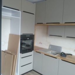 Lukasz Fabisiak - Jasnoszara zabudowa kuchenna z drewnianym blatem i czarnymi uchwytami. Widoczny piekarnik i zlew. Częściowo rozpakowane elementy meblowe w tle.