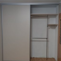 Lukasz Fabisiak - Jasna szafa wnękowa z przesuwnymi drzwiami i otwartą przestrzenią z półkami oraz drążkami na ubrania, widok na drewnianą podłogę.