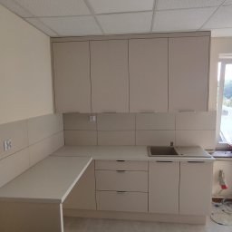 Lukasz Fabisiak - Jasna kuchnia na wymiar z górnymi szafkami i blatem roboczym, widok na zlew i fragment okna. Minimalistyczny design, neutralna kolorystyka, płytki na ścianie.