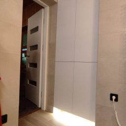 Lukasz Fabisiak - Biała szafa wnękowa z podświetleniem LED u dołu, widoczna z perspektywy wnętrza, z otwartymi drzwiami w tle. Minimalistyczny design, jasne płytki na ścianach i podłodze.