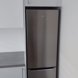 Lukasz Fabisiak - Nowoczesna kuchnia z zabudowaną lodówką Amica w srebrnej obudowie, otoczona białymi szafkami. Minimalistyczny design, szary blat. Górna szafka z uchwytem.