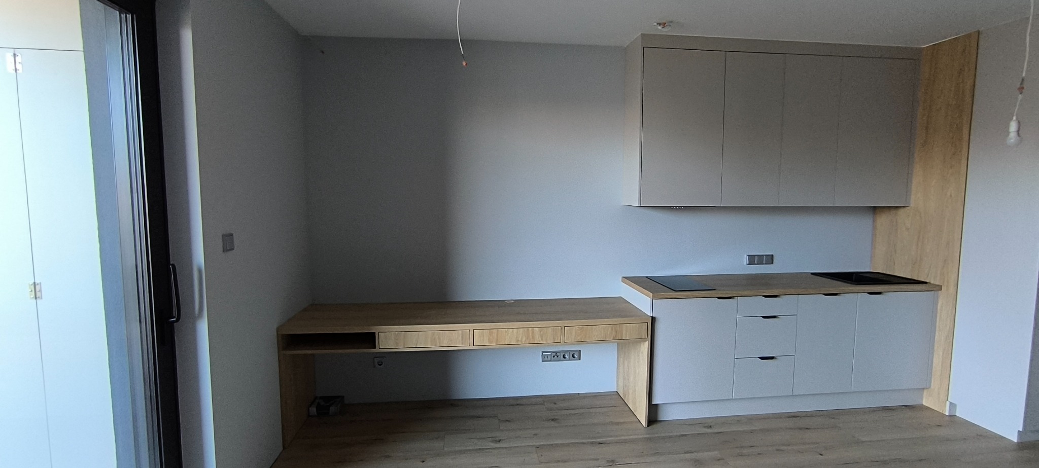 Jasna kuchnia na wymiar z drewnianym blatem i biurkiem w zabudowie, minimalistyczny design, szare fronty, drewniane akcenty, widok na podłogę i fragment okna.