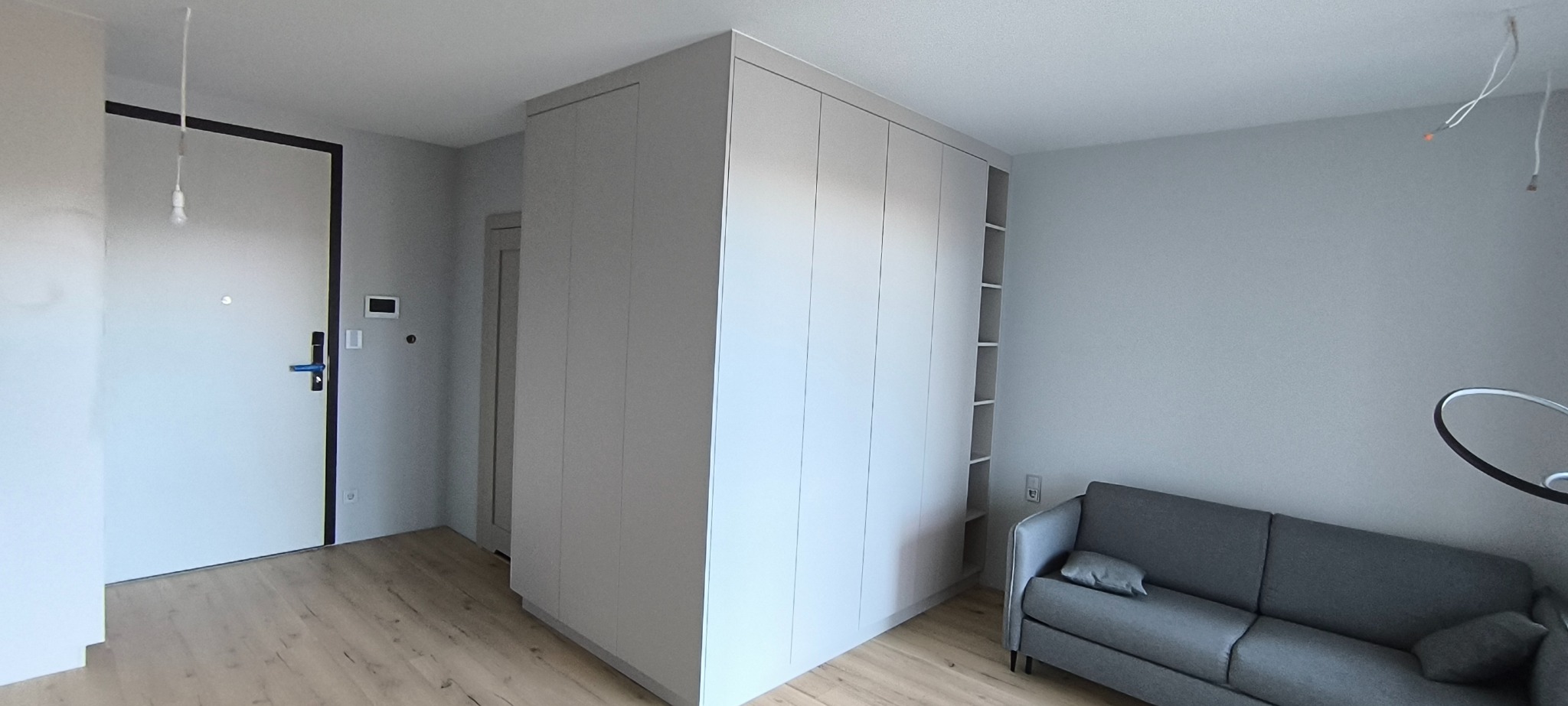 Jasnoszara szafa wnękowa z otwartymi półkami, dopasowana do koloru ścian, w nowoczesnym wnętrzu z szarą sofą i drewnianą podłogą.