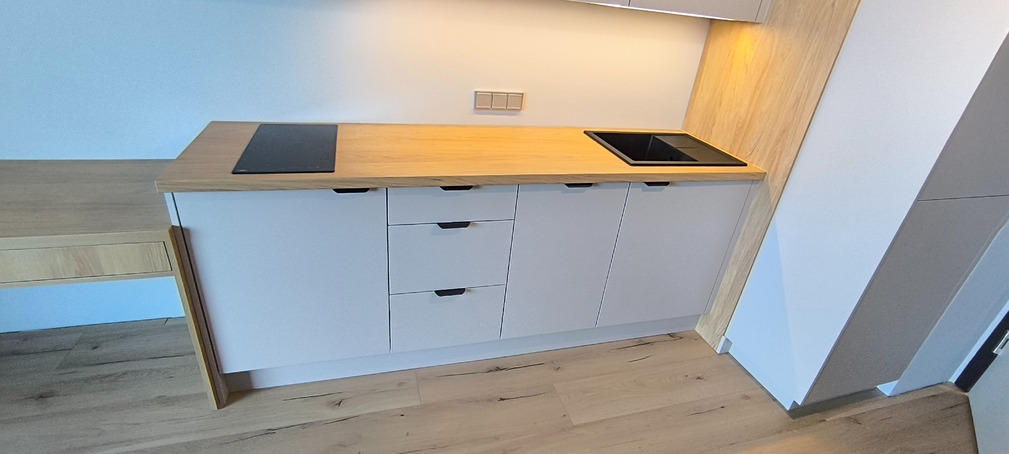 Jasna, minimalistyczna kuchnia z drewnianym blatem i białymi frontami. Widoczna płyta indukcyjna i zlew. Ujęcie z niskiego kąta podkreśla nowoczesny design.