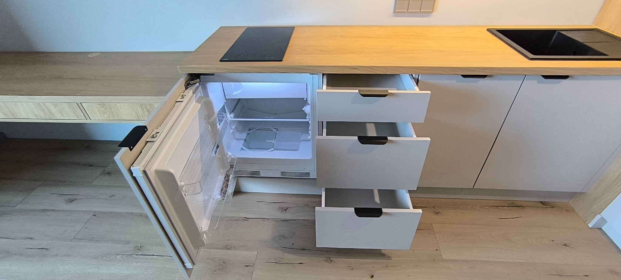 Jasna kuchnia z drewnianym blatem, płytą indukcyjną i zlewem. Otwarta lodówka w zabudowie i wysunięte szuflady. Nowoczesny design, minimalistyczne uchwyty.