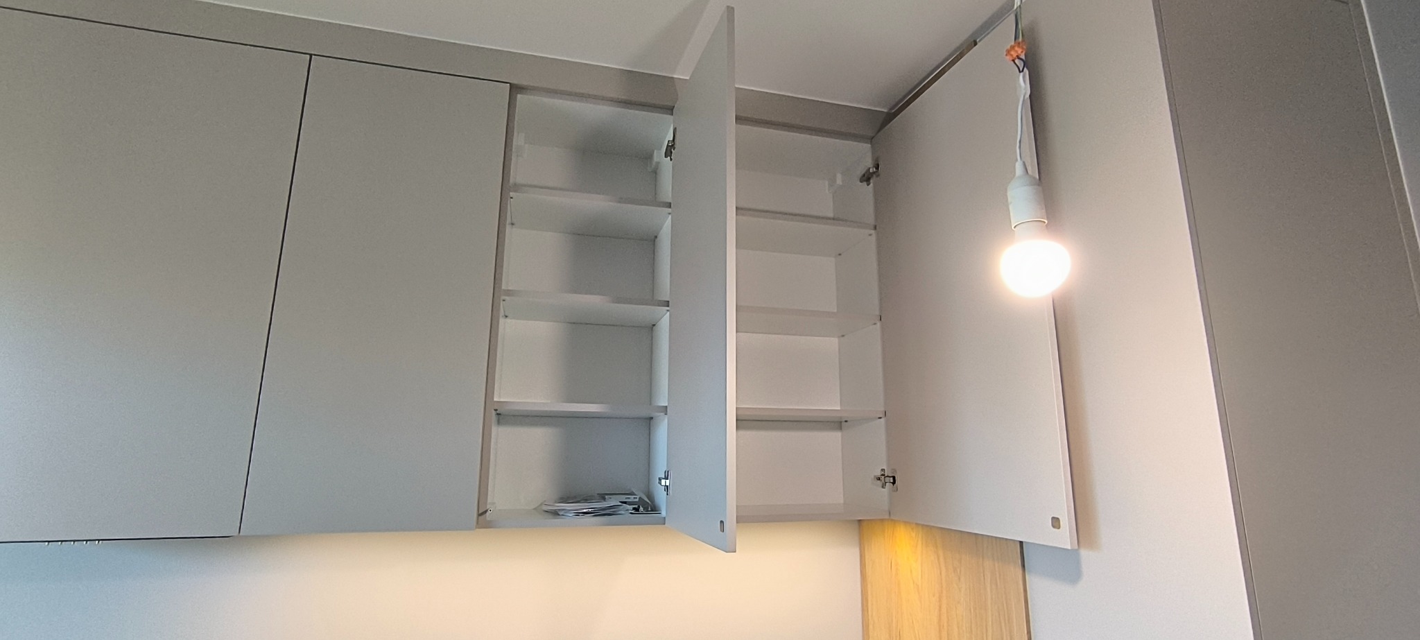 Szare szafki kuchenne wiszące z otwartymi drzwiczkami, widoczne półki wewnątrz. Oświetlenie punktowe pod szafkami i żarówka na kablu. Minimalistyczny design.
