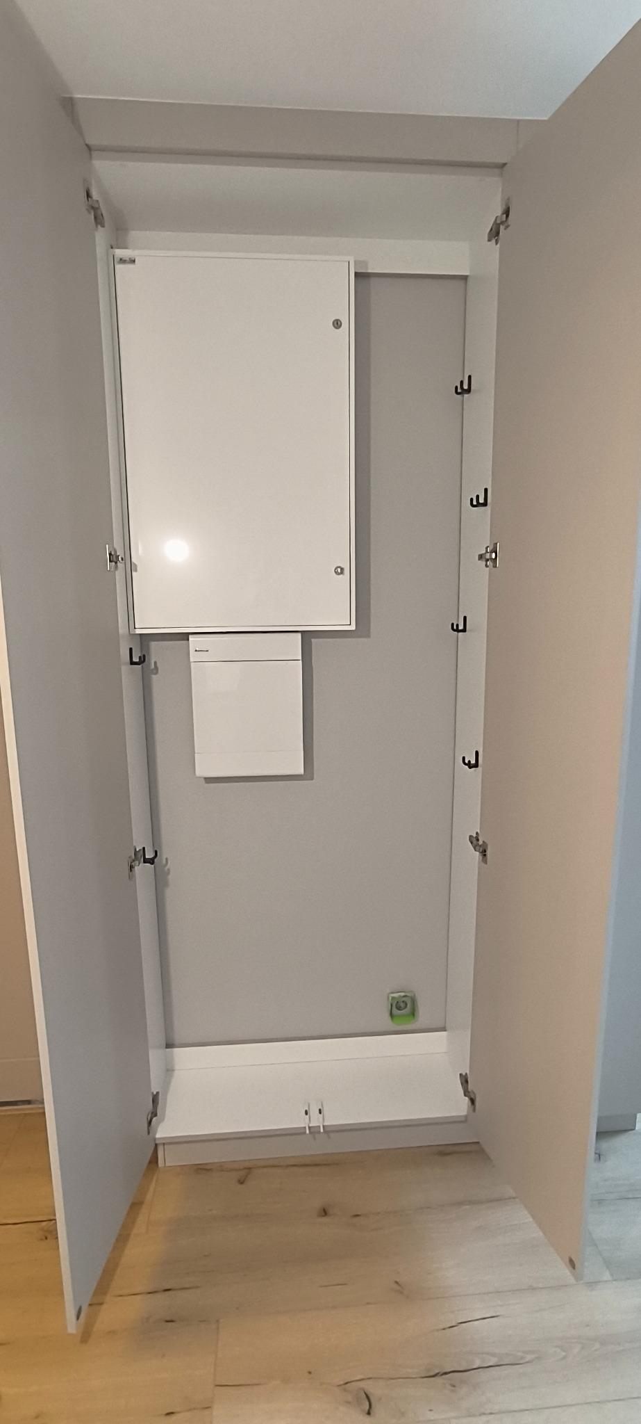 Otwarta szafa wnękowa z białymi drzwiami i haczykami na ścianie, z widoczną instalacją elektryczną wewnątrz. Minimalistyczny design, jasne wnętrze.