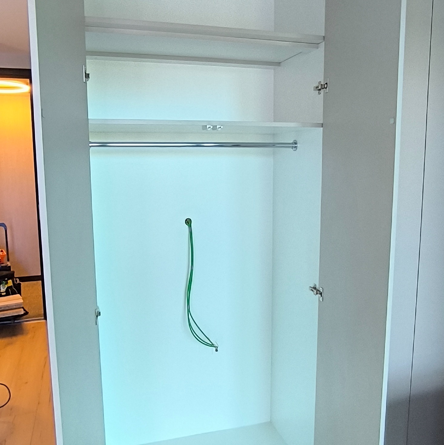 Wnętrze szafy wnękowej z drzwiami uchylnymi, widoczny drążek na ubrania i wystający kabel. Minimalistyczny design, jasne kolory.