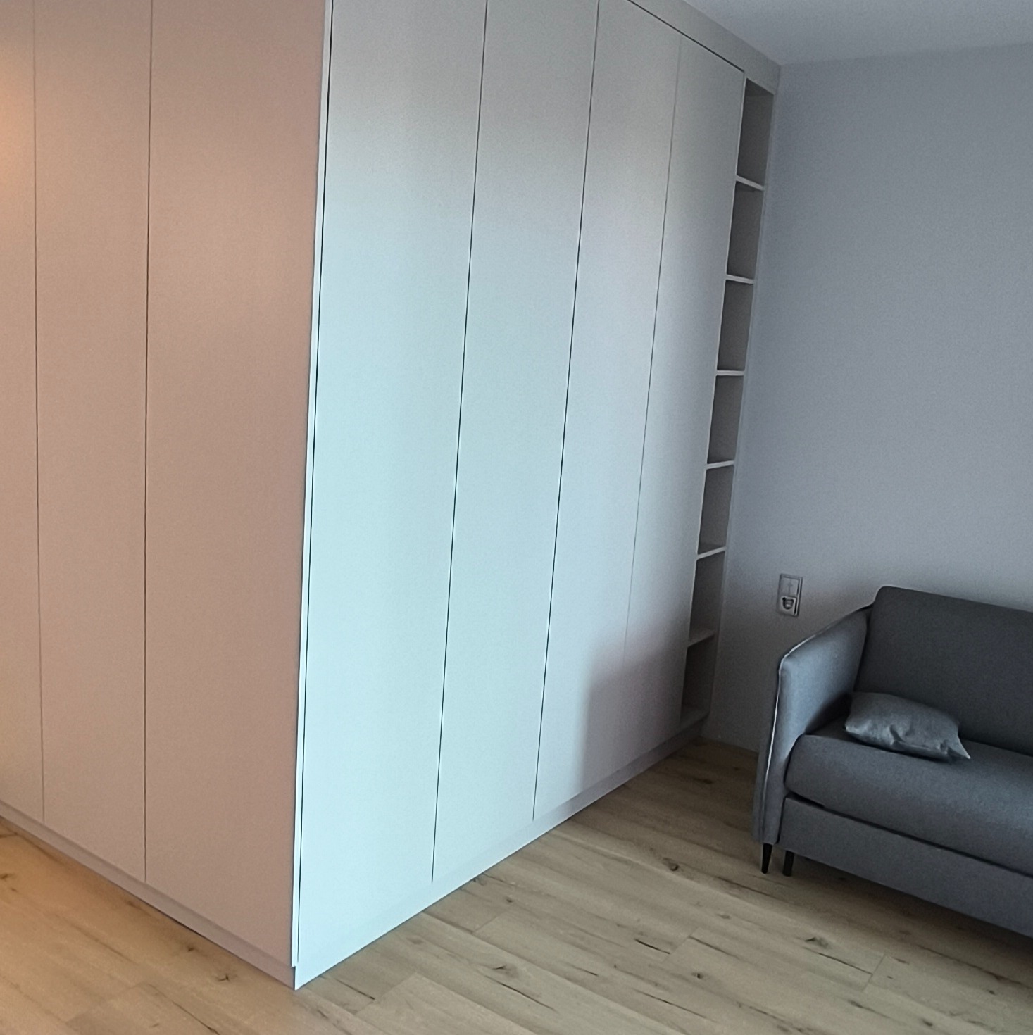 Jasna szafa wnękowa z otwartymi półkami, dopasowana do szarej sofy w minimalistycznym wnętrzu z drewnianą podłogą. Widok z boku, oświetlenie naturalne.