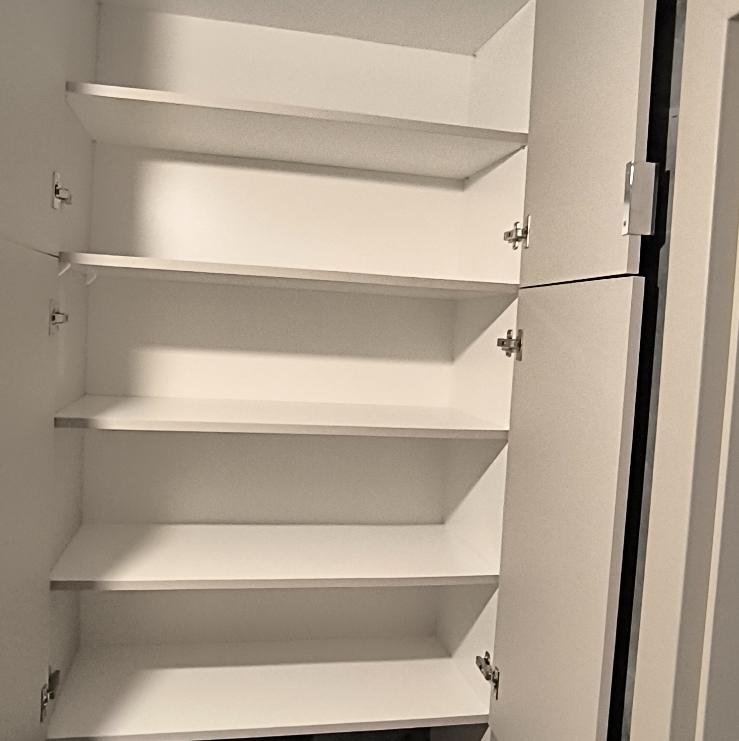 Otwarta, biała szafa z półkami, widok z bliska na wnętrze. Drzwiczki szafy częściowo otwarte, ukazujące metalowe zawiasy. Minimalistyczny design, jasne oświetlenie.