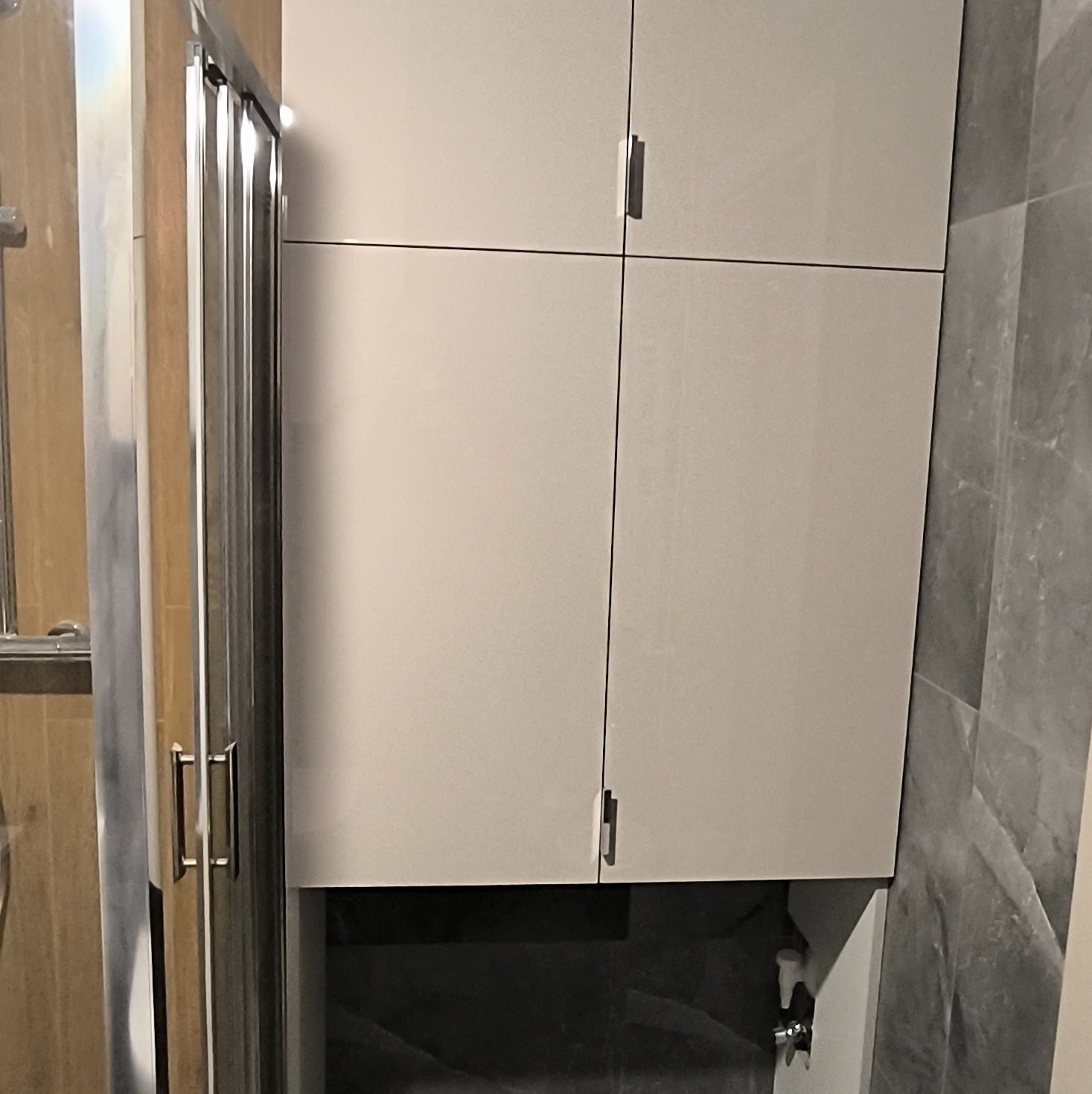 Jasna szafka łazienkowa z minimalistycznymi uchwytami, umieszczona nad umywalką, widoczna obok kabiny prysznicowej z chromowanymi elementami.