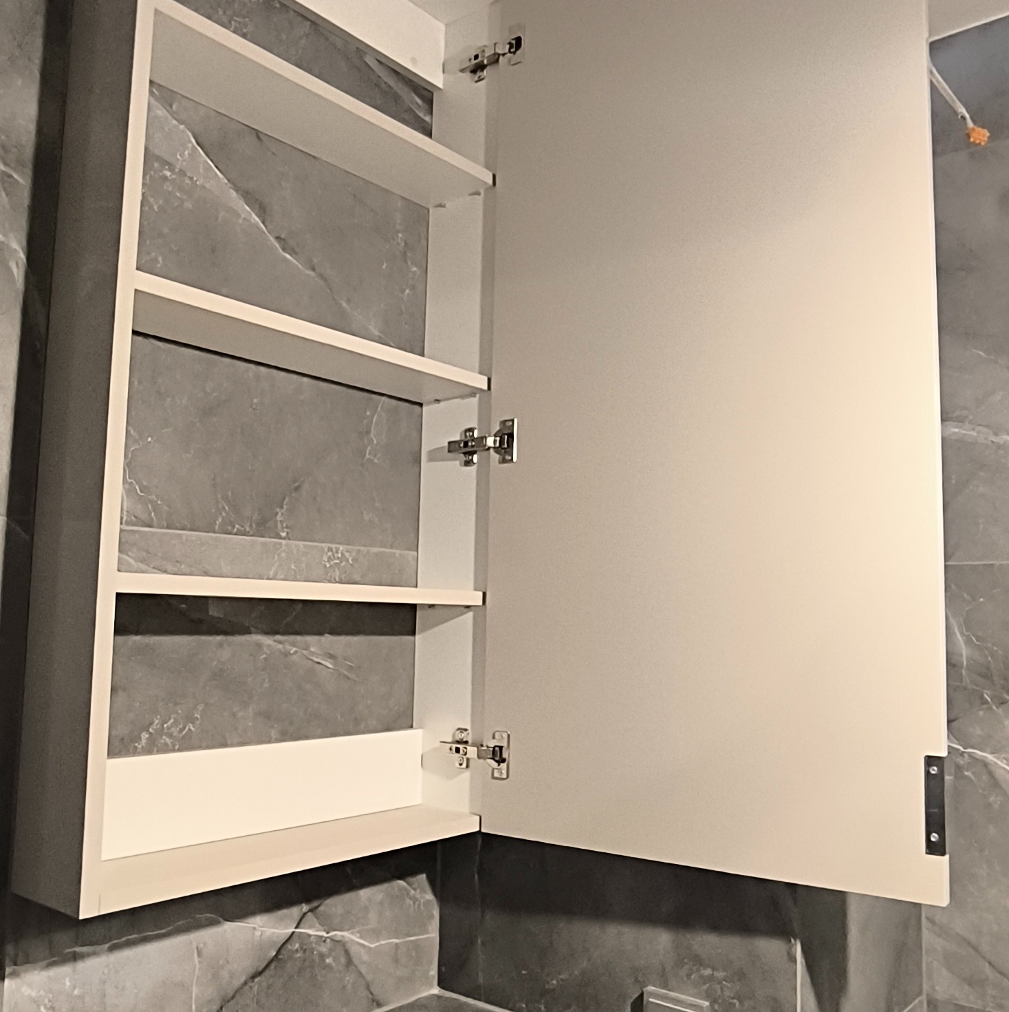Otwarta, biała szafka łazienkowa z półkami na tle szarej ściany imitującej marmur. Widoczne zawiasy i wnętrze szafki. Minimalistyczny design.
