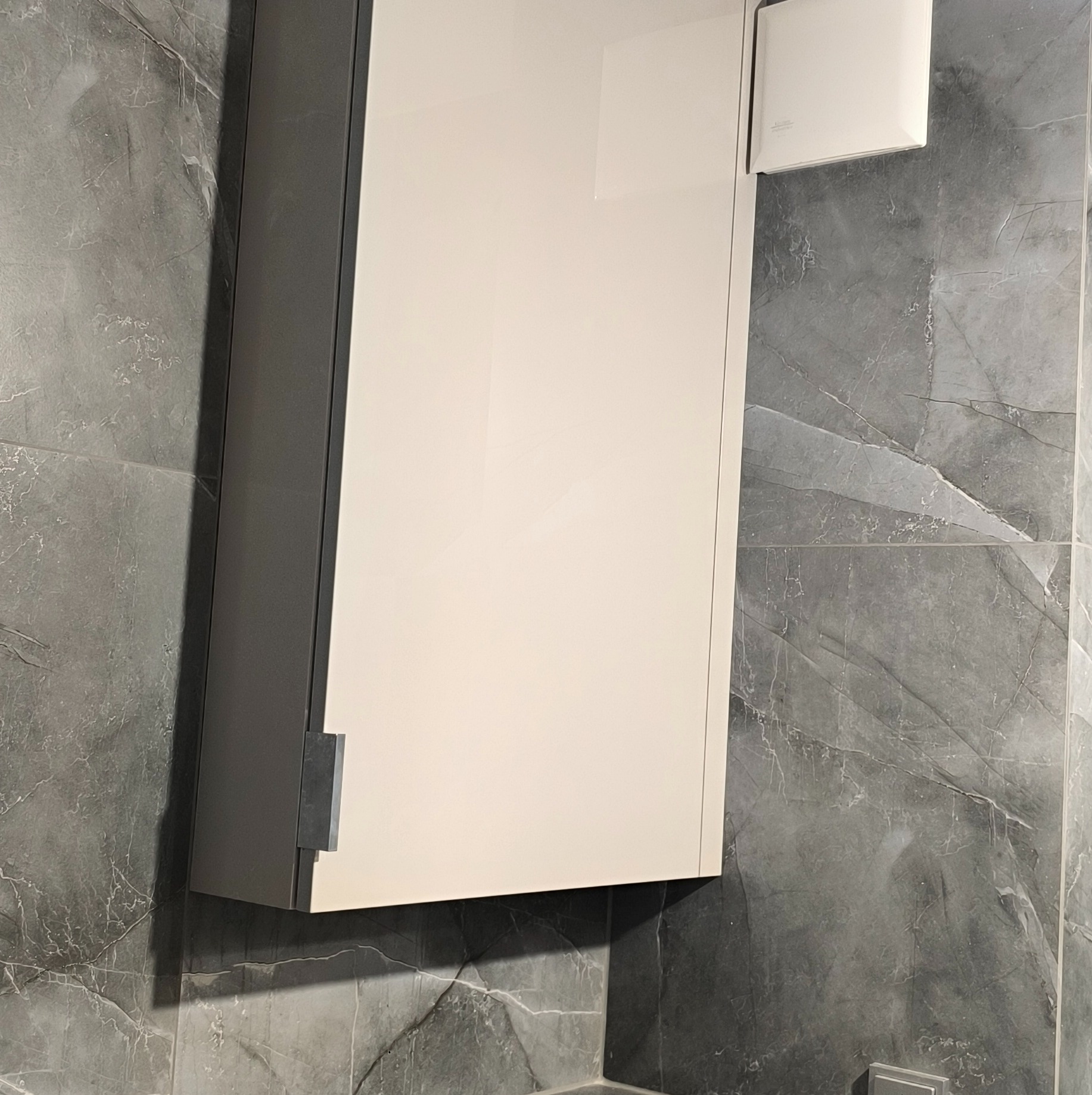 Biała szafka wisząca z szarymi bokami na tle ściany z szarego marmuru. Minimalistyczny design, nowoczesne wykończenie, widoczny fragment wentylatora.
