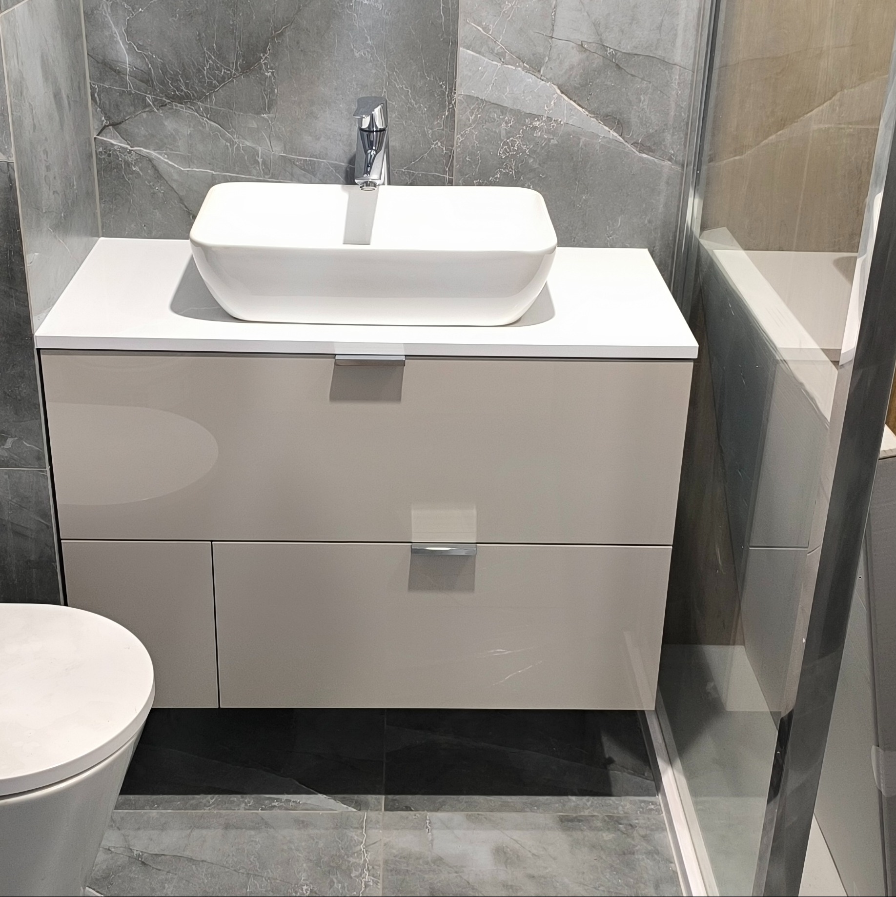 Beżowa szafka łazienkowa z umywalką nablatową na tle marmurowej ściany i kabiny prysznicowej. Minimalistyczny design, nowoczesne wykończenie.