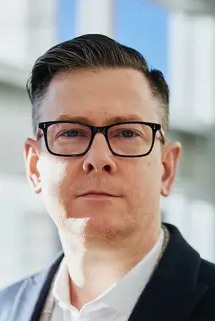 Portret mężczyzny w okularach i marynarce, na rozmytym tle biurowym.