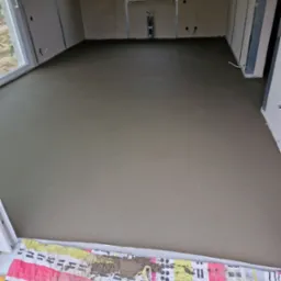 Świeżo wylana, gładka posadzka w surowym pomieszczeniu z oknem, zabezpieczona kolorową matą przy drzwiach balkonowych.