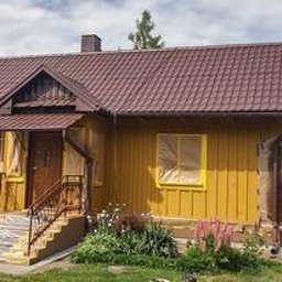 T&M Malowanie Dachów I Elewacji. - Żółta elewacja domu w trakcie malowania, okna zabezpieczone folią, brązowy dach z metalowej dachówki, widoczne kwiaty w ogrodzie przed domem.