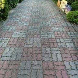 T&M Malowanie Dachów I Elewacji. - Częściowo wyczyszczona kostka brukowa w odcieniach szarości i czerwieni, widoczny efekt różnicy czystości, w tle zieleń krzewów.