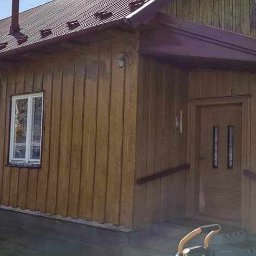 T&M Malowanie Dachów I Elewacji. - Drewniany dom z pomalowaną elewacją na brązowo, z metalowym dachem w kolorze burgundu, widok z zewnątrz w słoneczny dzień. Na pierwszym planie widoczny sprzęt do prac na wysokości.