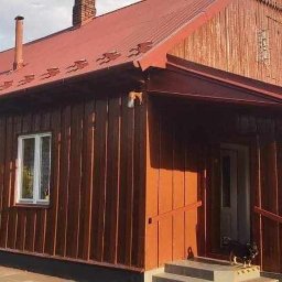 T&M Malowanie Dachów I Elewacji. - Czerwono-brązowy drewniany dom z czerwoną blachodachówką, widok z zewnątrz, mały pies na schodach wejściowych, w tle wieża kościelna.