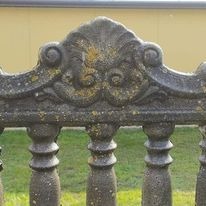 Szczegółowy widok ozdobnego, betonowego elementu balustrady z widocznymi śladami zabrudzeń i porostów, na tle trawnika i jasnej ściany budynku.