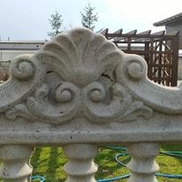 Dekoracyjny, betonowy element balustrady z ornamentem muszli i wolut na tle trawnika i pergoli.