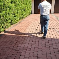 Pracownik w jeansach czyści czerwoną kostkę brukową na podjeździe za pomocą myjki ciśnieniowej. W tle zielony żywopłot i brama garażowa.