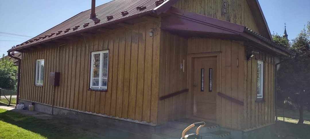 Drewniany dom z pomalowaną elewacją na brązowo, z metalowym dachem w kolorze burgundu, widok z zewnątrz w słoneczny dzień. Na pierwszym planie widoczny sprzęt do prac na wysokości.